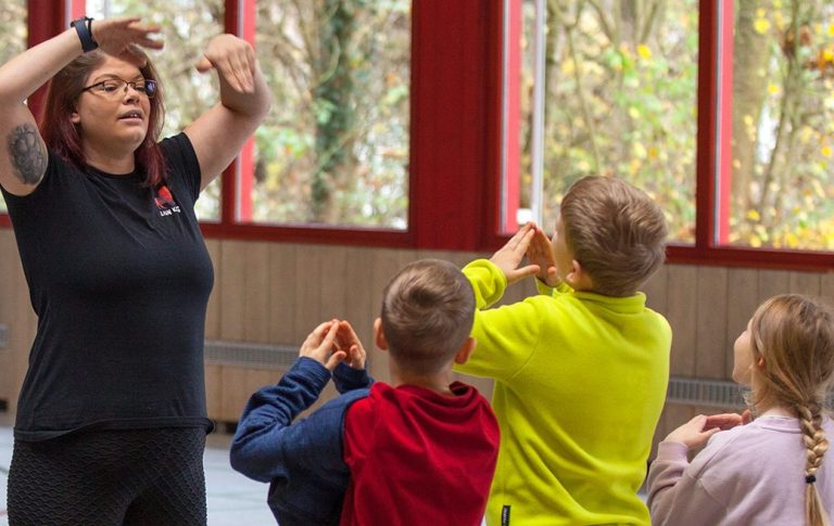 lion-kids-resilienzkurs1.jpg Die Resilienztrainerin Helen Häge zeigt einer Gruppe von Kindern Bewegungen in einem Raum mit großen Fenstern.